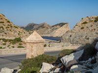 two stone walls next to a road near a mountain and water in the distance there is a small bridge