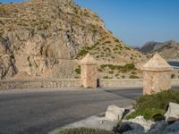 two stone walls next to a road near a mountain and water in the distance there is a small bridge