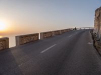 a paved road passing by a cliff near the ocean at sunset with people sitting on a ledge in the distance