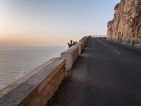 a paved road passing by a cliff near the ocean at sunset with people sitting on a ledge in the distance