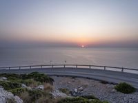 the sunset is rising over the water and the road next to it is paved by a curvy, curved fence