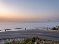 the sunset is rising over the water and the road next to it is paved by a curvy, curved fence