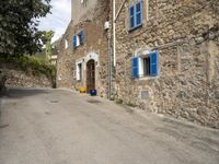 an empty street with blue shutters on two windows and stone walls behind a building