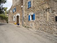 an empty street with blue shutters on two windows and stone walls behind a building