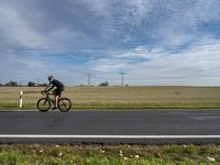 man in bicycle helmet on the road looking down at something in the distance with an empty field behind him