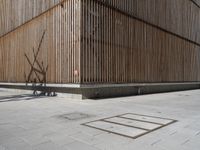 a man riding his bicycle on a concrete sidewalk outside of a building made out of wood planks