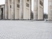 a man sits on a bench in an empty square between large stone pillars on top of snow covered ground