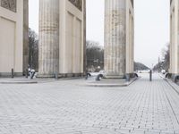 a man sits on a bench in an empty square between large stone pillars on top of snow covered ground