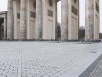 a man sits on a bench in an empty square between large stone pillars on top of snow covered ground