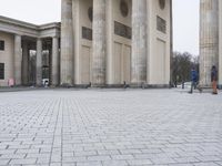a man sits on a bench in an empty square between large stone pillars on top of snow covered ground