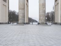 a man sits on a bench in an empty square between large stone pillars on top of snow covered ground