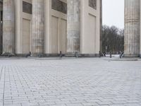 a man sits on a bench in an empty square between large stone pillars on top of snow covered ground