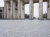 a man sits on a bench in an empty square between large stone pillars on top of snow covered ground