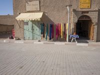 the man is sitting in front of his shop in the city street, while holding his hands to his knee