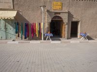 the man is sitting in front of his shop in the city street, while holding his hands to his knee