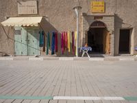 the man is sitting in front of his shop in the city street, while holding his hands to his knee