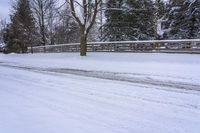 a man snow skiing down a snow covered road by a wooded area next to a fence