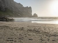 there is a man standing on the beach near some rocks and water behind him are a lot of footprints