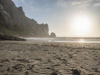 there is a man standing on the beach near some rocks and water behind him are a lot of footprints