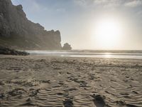 there is a man standing on the beach near some rocks and water behind him are a lot of footprints