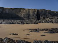 a man standing on top of a sandy beach under a cliff next to the ocean