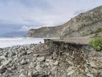 there is a man with a surfboard on a rocky beach by the ocean and mountains