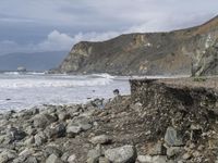 there is a man with a surfboard on a rocky beach by the ocean and mountains