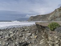 there is a man with a surfboard on a rocky beach by the ocean and mountains