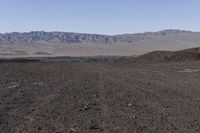 a man walking on a field in a desert land with a motorcycle behind him and a mountain in the distance