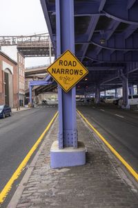 there is a road narrows sign under a bridge above it on a sidewalk in the city