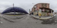 a fisheye image of a bus stop and pedestrian bridge over a street in front of a large brick building