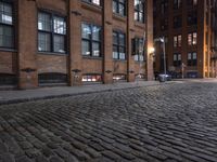 a row of old brick buildings at night on a cobble stone street with street lamps