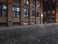 a row of old brick buildings at night on a cobble stone street with street lamps