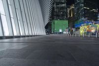 an empty sidewalk with many benches and some skyscrapers in the background at nighttime