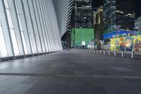 an empty sidewalk with many benches and some skyscrapers in the background at nighttime