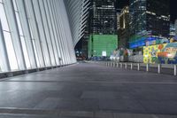 an empty sidewalk with many benches and some skyscrapers in the background at nighttime