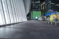 an empty sidewalk with many benches and some skyscrapers in the background at nighttime