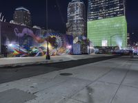 a night view with skyscrapers in the background and a painted wall next to the sidewalk