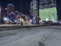 a night view with skyscrapers in the background and a painted wall next to the sidewalk