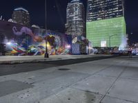 a night view with skyscrapers in the background and a painted wall next to the sidewalk