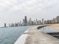 a long concrete pier has water on it near the city skyline on the ocean and skyscrapers