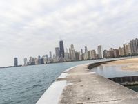 a long concrete pier has water on it near the city skyline on the ocean and skyscrapers