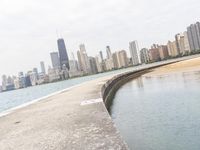 a long concrete pier has water on it near the city skyline on the ocean and skyscrapers