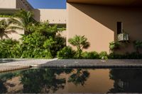 an empty pool in front of a tan building with small windows on it, and lush vegetation around