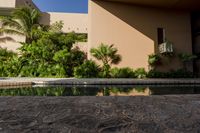 an empty pool in front of a tan building with small windows on it, and lush vegetation around