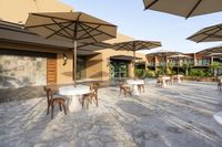 the courtyard has many umbrellas and tables with chairs with people seated at them and there is a large pool in the background