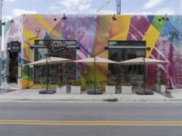 several umbrellas and benches are outside a restaurant in the city with multicolored mural