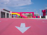 a parking lot with an arrow and a pink building in the background, with buildings and a walkway in the middle