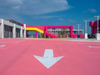 a parking lot with an arrow and a pink building in the background, with buildings and a walkway in the middle
