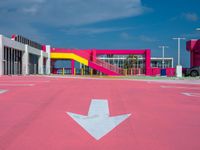 a parking lot with an arrow and a pink building in the background, with buildings and a walkway in the middle